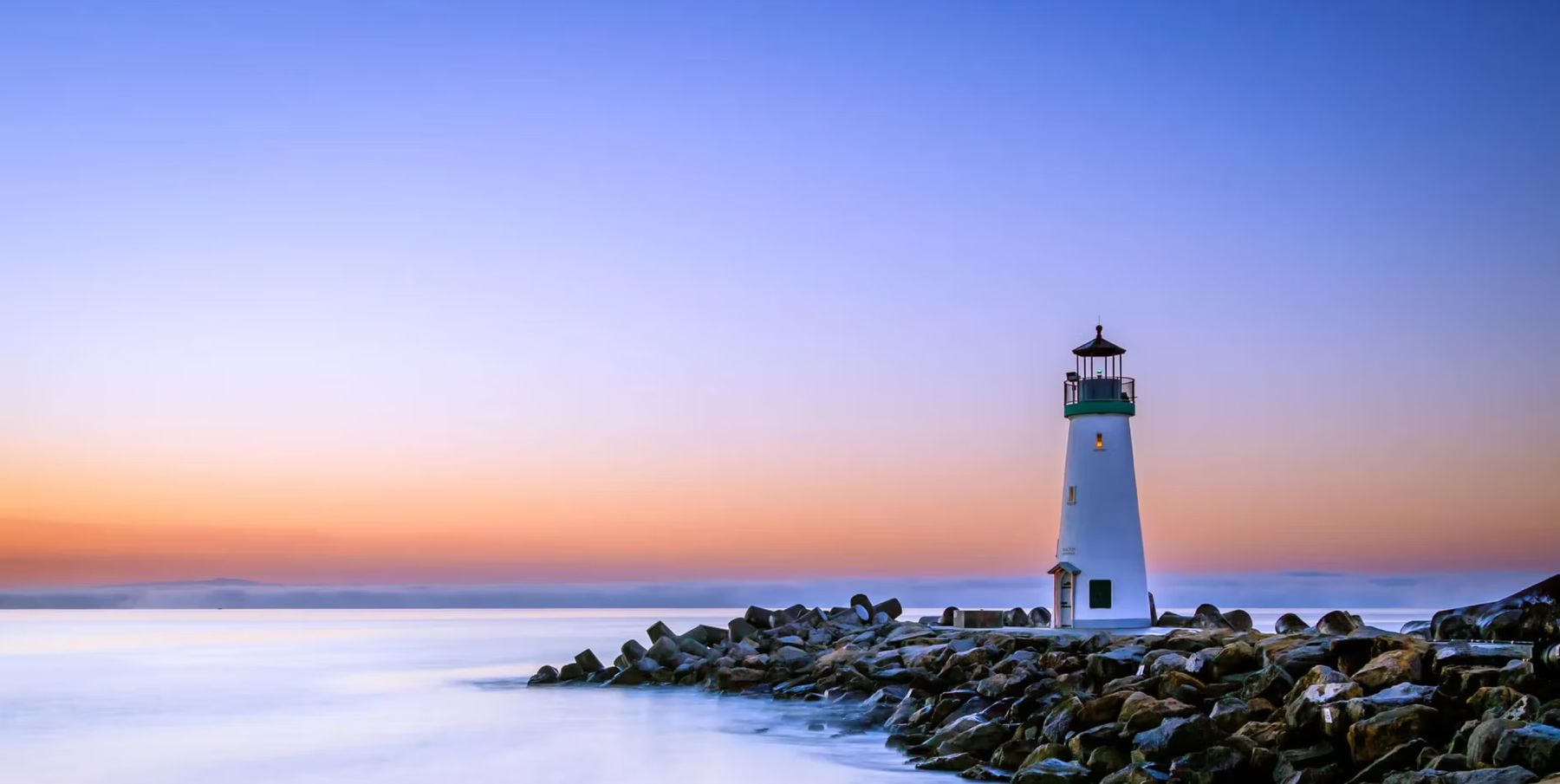 White lighthouse on rocky shore at sunset with calm sea and colorful sky.